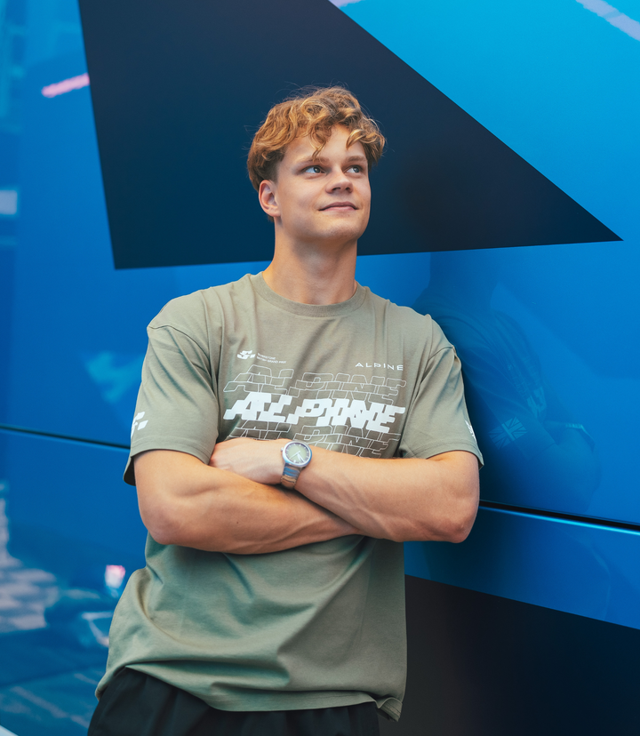 Young man wearing olive green Alpine T shirt with large white graphic across chest against a blue background