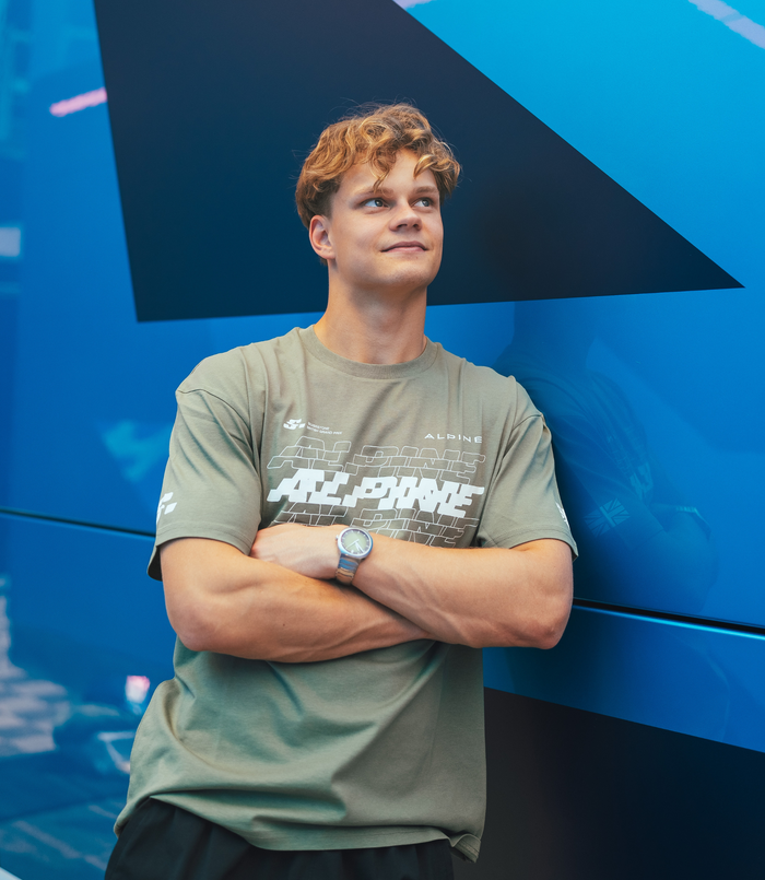 Young man wearing olive green Alpine T shirt with large white graphic across chest against a blue background