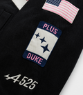 Close-up of patches on a dark racing jacket including a pink striped flag a blue patch with DUKE lettering and stars and a US flag badge