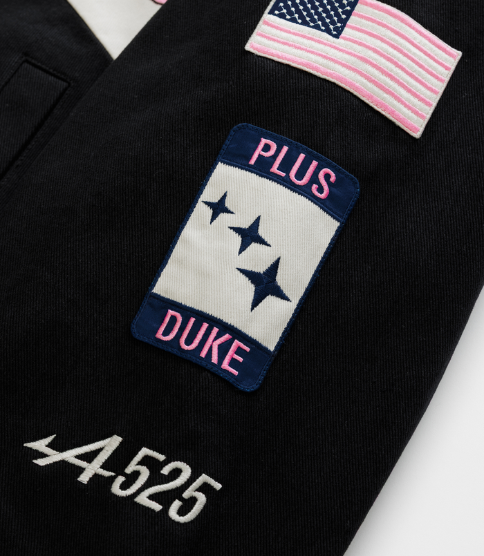 Close-up of patches on a dark racing jacket including a pink striped flag a blue patch with DUKE lettering and stars and a US flag badge