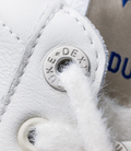 Close-up of a white Duke + Dexter Plus Duke sneaker showing a circular metal eyelet, textured leather, and fluffy trim.