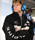 Side profile of a man in a black Duke and Dexter racing jacket wearing a blue over-ear headset in a motorsport workshop