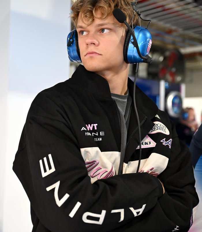 Side profile of a man in a black Duke and Dexter racing jacket wearing a blue over-ear headset in a motorsport workshop
