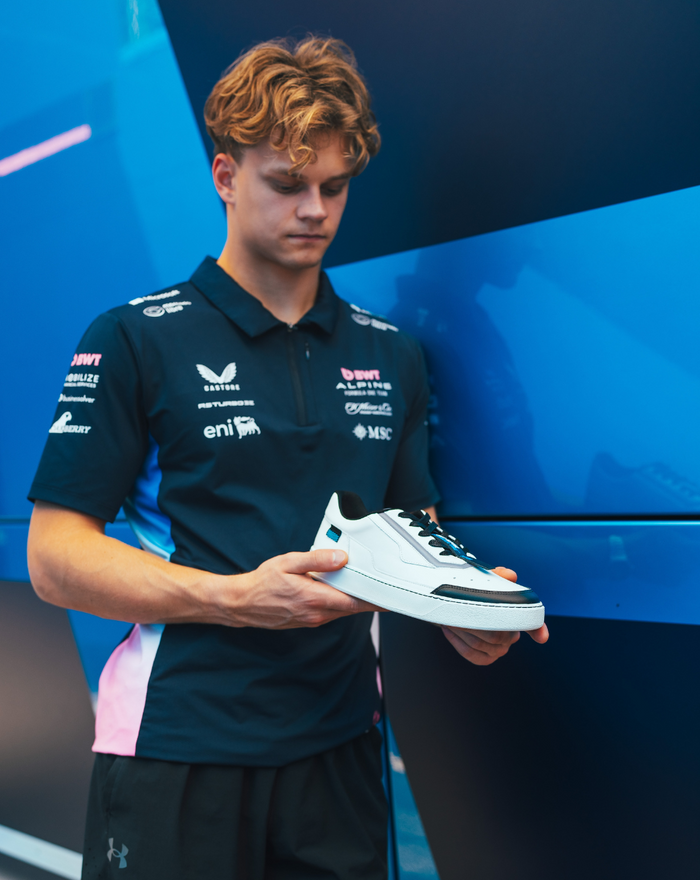 Young man in a BWT Alpine team polo holds a white Endstate sneaker against a blue backdrop.