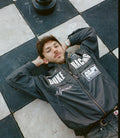 Man wearing a black Duke & Dexter trackside jacket lying on checkered pavement with large white lettering on the chest.