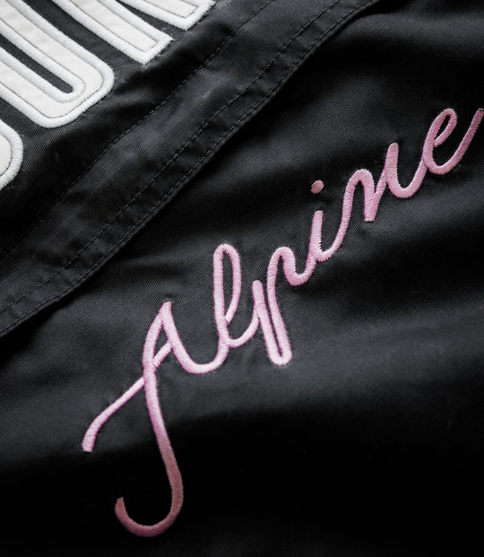 Close-up of black trackside jacket showing white embroidered patch lettering and pink cursive script