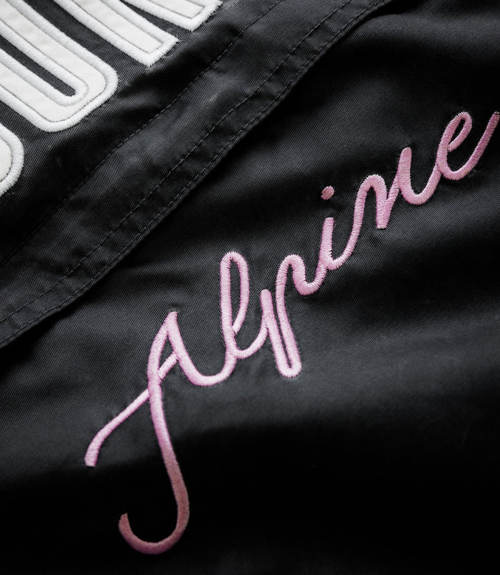 Close-up of black trackside jacket showing white embroidered patch lettering and pink cursive script
