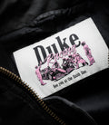 Close up of a white patch on black fabric showing Duke & Dexter branding in black and pink with the finish line slogan