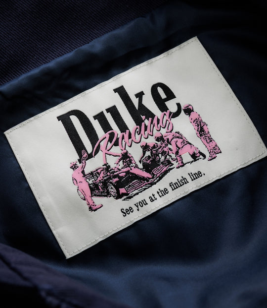 White stitched label on navy fabric with pink racing graphics and the Duke & Dexter branding