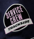 Close up of a circular embroidered patch on navy fabric with pink lettering and a white border.