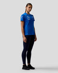 Full-body view of a woman wearing a blue BWT Alpine Driver Set Up T Shirt with team logos on the chest and sleeve