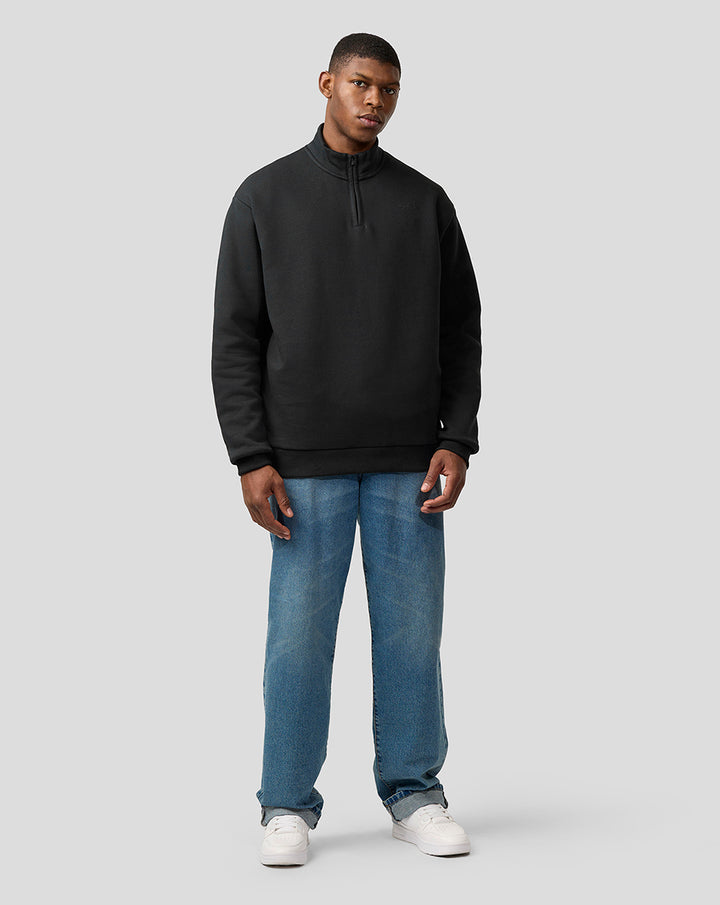 Male model wearing black oversized Alpine Castore quarter zip pullover with funnel neck.