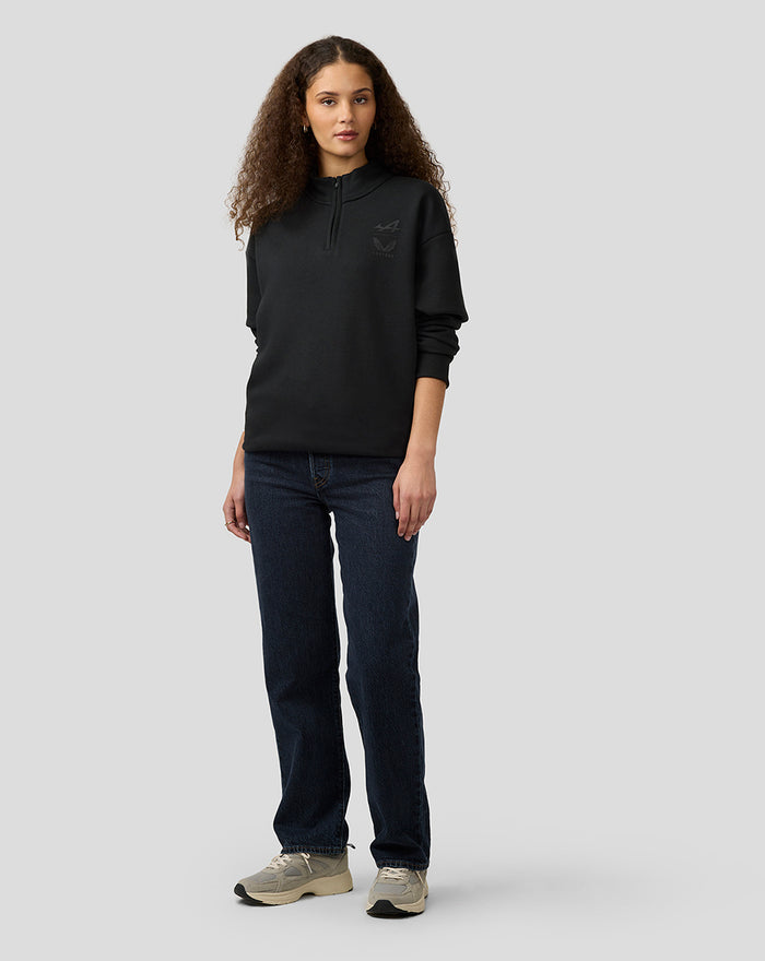 Female model in black oversized Alpine Castore quarter zip pullover with funnel neck and chest logos.