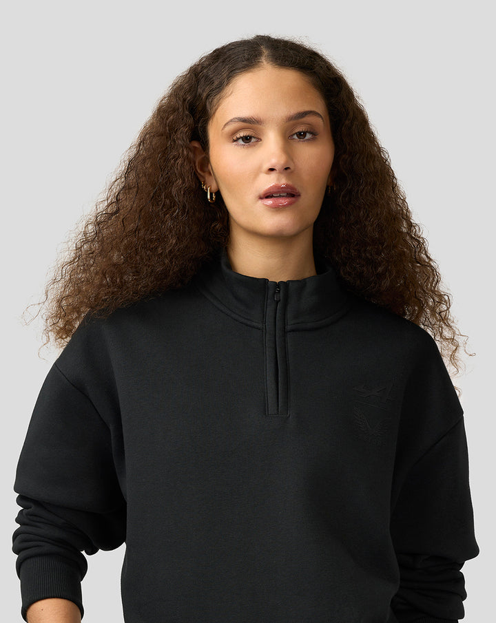 Woman wearing black Alpine Castore funnel-neck quarter-zip fleece with chest logo.